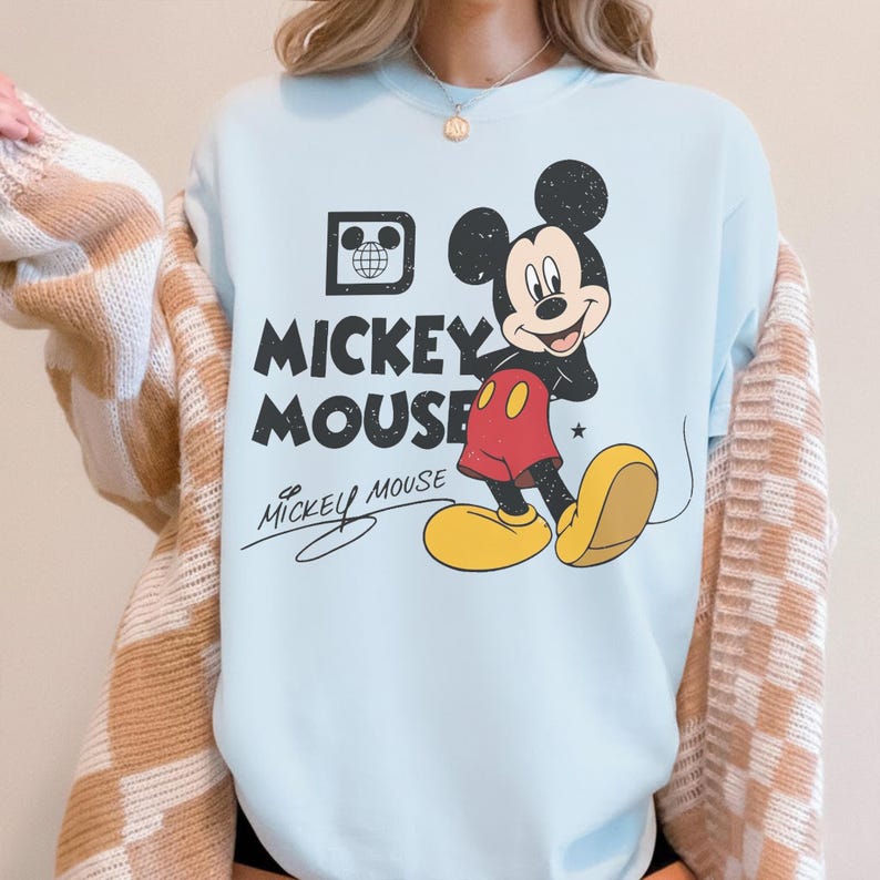 May include: Light blue sweatshirt featuring a graphic of Mickey Mouse. The design includes Mickey in his classic outfit, with the words "Mickey Mouse" in bold letters. A small graphic of a globe is also present.