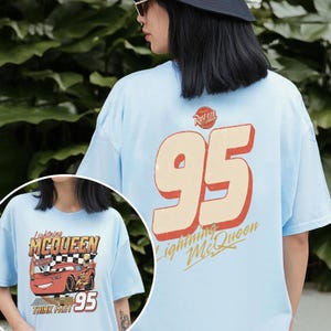 Op de afbeelding: Lichtblauw T-shirt met Lightning McQueen uit de film Cars. De voorkant toont de auto met de tekst "Lightning McQueen", "Think Fast" en "95". De achterkant heeft een grote "95" met "Lightning McQueen" en "Rusteze".