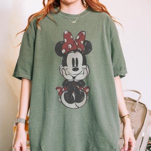 May include: An olive green t-shirt featuring a distressed graphic of Minnie Mouse. Minnie is depicted with a red polka dot bow and matching shoes. The shirt has short sleeves and a relaxed fit, suitable for casual wear.