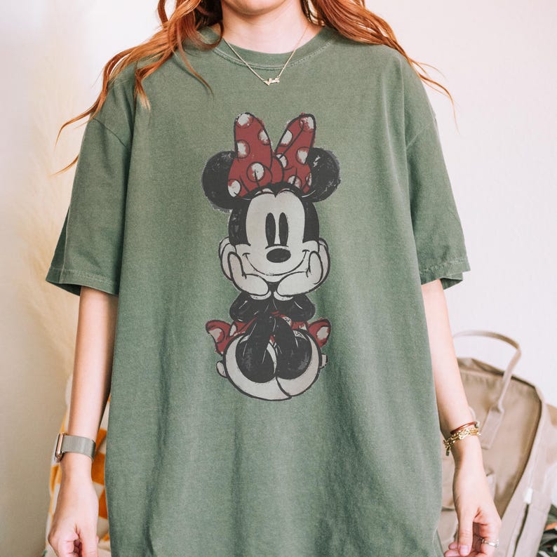 May include: An olive green t-shirt featuring a distressed graphic of Minnie Mouse. Minnie is depicted with a red polka dot bow and matching shoes. The shirt has short sleeves and a relaxed fit, suitable for casual wear.