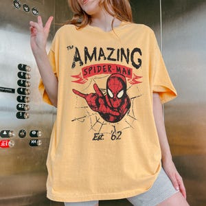 May include: Mustard yellow t-shirt with a Spider-Man graphic. The design includes the text "The Amazing Spider-Man" and "Est. '62", along with a red Spider-Man figure and a spider web. The shirt has short sleeves.