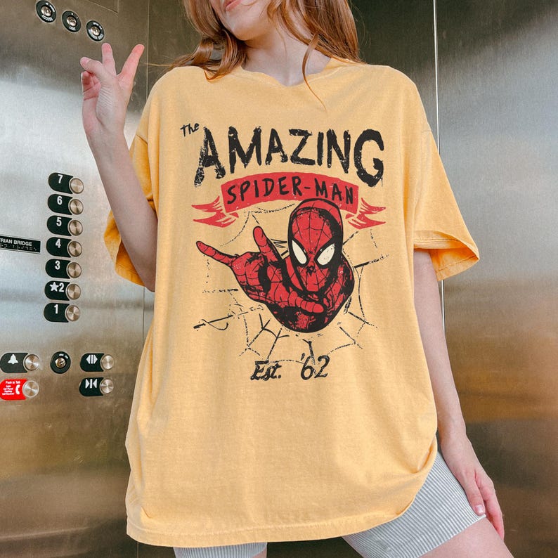 May include: Mustard yellow t-shirt with a Spider-Man graphic. The design includes the text "The Amazing Spider-Man" and "Est. '62", along with a red Spider-Man figure and a spider web. The shirt has short sleeves.