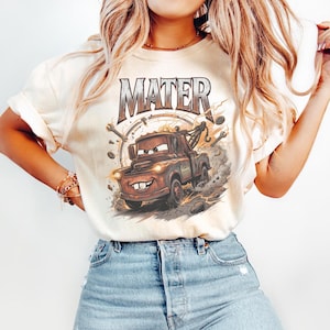 Pu&ograve; includere: T-shirt color crema con una grafica del personaggio Mater del film Cars. Il design include il nome "MATER" in un font invecchiato sopra un'illustrazione del carro attrezzi. La grafica &egrave; circondata da strumenti e fulmini.
