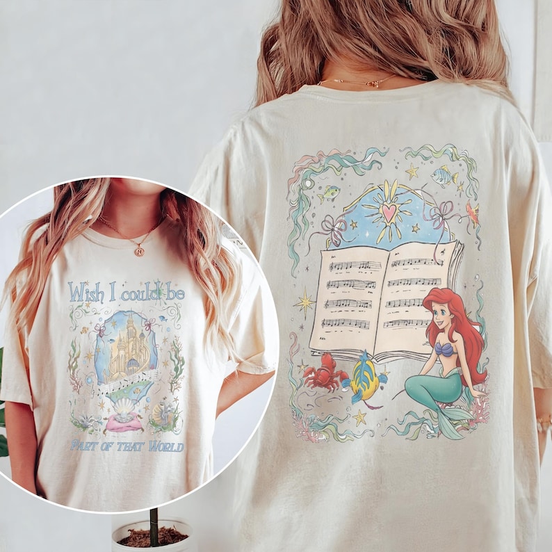 May include: Cream-coloured t-shirt with a colourful illustration of a mermaid, musical notes, and the text "Wish I could be part of that world." The design includes a castle, sea creatures, and a heart.