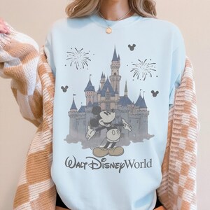 Puede incluir: Sudadera azul claro con un gr&aacute;fico de Mickey Mouse frente al castillo de Cenicienta en Walt Disney World. El dise&ntilde;o incluye fuegos artificiales y siluetas de Mickey Mouse. El texto "Walt Disney World" est&aacute; impreso debajo.