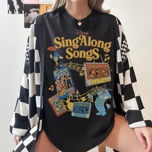 Puede incluir: Camiseta negra con estampado de "Disney Sing Along Songs" con varios personajes de Disney y cintas de cassette. La camiseta se combina con una rebeca a cuadros en blanco y negro.
