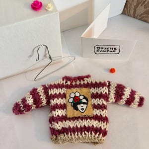 May include: A small, hand-knitted sweater with a striped pattern in burgundy and white. The sweater has a patch on the front with a cartoon face and a flower.