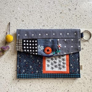 Peut inclure: Un portefeuille patchwork avec un porte-clés. Le portefeuille présente une variété de tissus, notamment des pois, des imprimés floraux et un motif pied-de-poule. Il a une fermeture à bouton orange et un petit détail brodé.