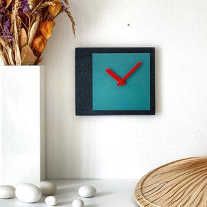 Peut inclure: Une horloge carrée de couleur bleu-vert et noire avec des aiguilles rouges sur un mur blanc. L'horloge est suspendue au-dessus d'un vase blanc avec des fleurs séchées.