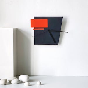 Puede incluir: Un reloj de pared moderno con una esfera negra y detalles naranjas. Las manecillas del reloj son negras y la esfera está ligeramente inclinada.