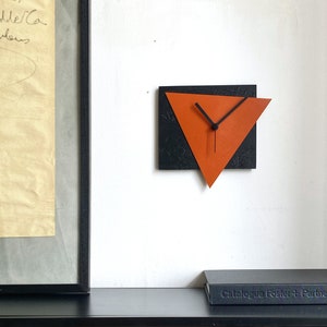 May include: A modern wall clock with a black background and an orange triangle shape with black hands. The clock is mounted on a white wall.
