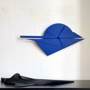 Puede incluir: Un reloj de pared geométrico azul con un diseño único. La esfera del reloj es un semicírculo con una aguja larga y fina y una aguja más corta y gruesa.