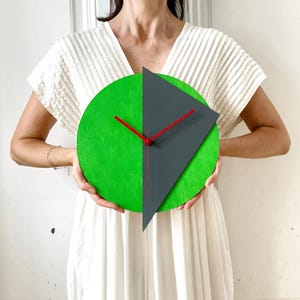 Pode incluir: Um relógio circular verde e cinza com ponteiros vermelhos. O relógio tem um design geométrico com uma forma triangular do lado direito.