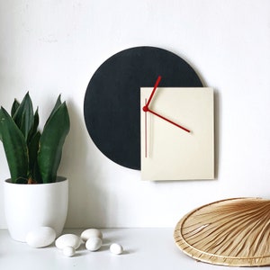Könnte beinhalten: Eine moderne Wanduhr mit schwarzem runden Hintergrund und einem weißen quadratischen Zifferblatt mit roten Zeigern. Die Uhr ist an einer weißen Wand montiert.