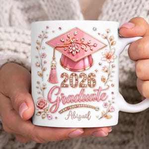 Może przedstawiać: Biały ceramiczny kubek z motywem ukończenia szkoły, z różową czapką absolwenta, rokiem 2026 i napisem "Graduate" w ozdobnej czcionce. Ozdoby kwiatowe i imię "Abigail" również widoczne. Idealny prezent dla absolwenta.