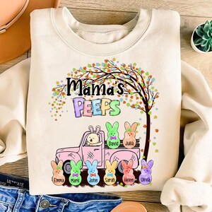Peut inclure: Un sweat-shirt blanc avec un motif de camion rose transportant une famille de lapins aux couleurs pastel. Les lapins sont &eacute;tiquet&eacute;s avec des noms comme David, Julia, Emma, Mark, John, Sarah, Helen et Olivia. Le texte "Mama's Peeps" est au-dessus du camion.