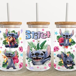 May include: Three clear glass tumblers with bamboo lids and metal straws. Each tumbler features a colourful design of the character Stitch from Lilo & Stitch, surrounded by tropical flowers and foliage. The word "Stitch" is printed on one tumbler.