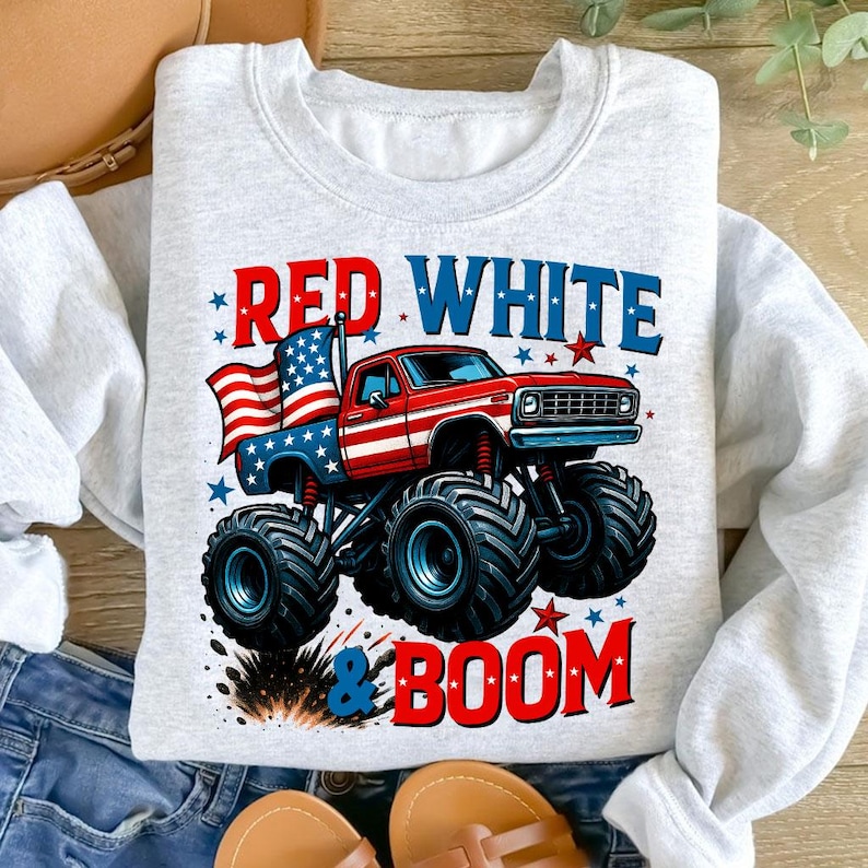 May include: A light gray sweatshirt featuring a red, white, and blue monster truck design. The truck has an American flag in the bed and the words "RED WHITE & BOOM" in bold letters. The design includes stars and a burst effect.