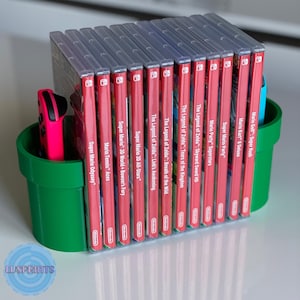 May include: A green and black video game console stand with two slots for controllers and a shelf for storing video game cartridges. The shelf is filled with 12 video game cartridges, including titles like "Super Mario Odyssey", "Mario Tennis Aces", and "The Legend of Zelda: Breath of the Wild".