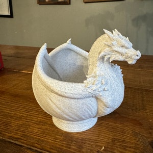 Op de afbeelding: Een witte keramische plantenbak in de vorm van een draak met zijn kop naar de zijkant gedraaid. De plantenbak heeft een gespikkelde textuur en een brede, open mond.