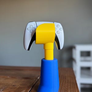 May include: A white and black video game controller is held in a yellow and blue 3D printed stand. The stand has a yellow base and a blue top.