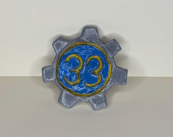 May include: A gray gear-shaped object with a blue center. The number "33" is written in gold within the blue circle. The gear has a textured appearance, and the background is a neutral color.