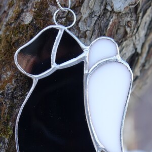 May include: A stained glass ornament depicting a black figure with a white wing. The figure is wearing a long, flowing robe and has a halo. The ornament is made of glass and has a silver wire frame.