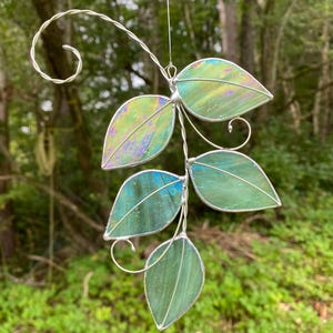 May include: A handcrafted stained glass leaf mobile with five iridescent green leaves. The leaves are connected by a silver wire stem and a decorative swirl at the top. The background is a blurred view of a forest.