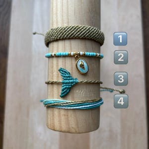 May include: Four handmade bracelets in gold, teal, and white are displayed on a wooden dowel. One bracelet is woven gold, another features turquoise beads and a shell charm, and a third has a teal macrame mermaid tail.
