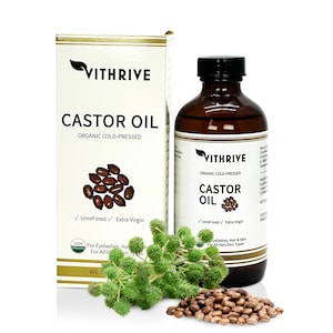 May include: A brown glass bottle of Vithrive Organic Cold-Pressed Castor Oil. The bottle is labeled with the product name and ingredients. The bottle is sitting on a white surface with castor beans and a castor plant.