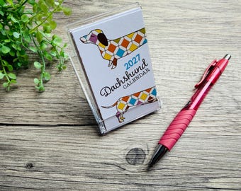 May include: A 2027 Dachshund calendar in a clear plastic stand featuring a colorful geometric dog illustration and the text 2027 Dachshund Calendar, accompanied by a retractable red ballpoint pen with a textured grip on the barrel.