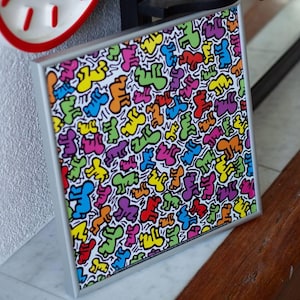 Puede incluir: Una obra de arte enmarcada con un patrón de figuras coloridas en azul, verde, amarillo, naranja y morado sobre un fondo blanco. La obra está en un marco plateado y descansa sobre una superficie de madera.