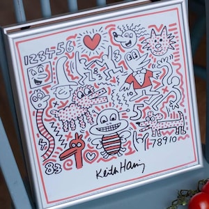 May include: A square framed print featuring a white background with black line art and coral-colored accents. The artwork displays various cartoon characters, numbers, and symbols, including a heart and a bee. The signature Keith Haring is printed at the bottom.
