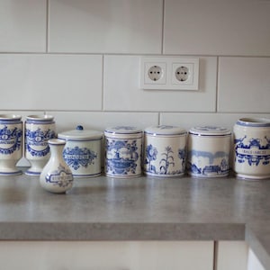 Może przedstawiać: Kolekcja niebiesko-białych ceramicznych pojemników kuchennych i wazonów ustawiona na blacie. Pojemniki mają pokrywki i posiadają różne wzory dekoracyjne. Tło obejmuje białe płytki i podwójne gniazdo elektryczne.