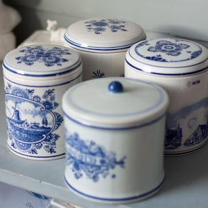 May include: A collection of white ceramic jars with blue floral and landscape designs. Each jar has a matching lid, and the designs vary, including windmills and stylized flowers. The jars are arranged on a light blue surface.