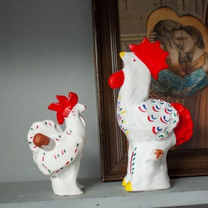 May include: Two white ceramic rooster figurines with bright red combs and wattles. The bodies are decorated with small, repeating crescent-shaped patterns in red, blue, and green. The larger rooster has yellow feet, while both feature detailed, hand-painted markings.