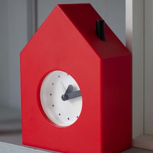 Op de afbeelding: Een felrode tafelklok in de vorm van een huis met een witte ronde wijzerplaat. De klok heeft zwarte uur- en minuutwijzers, zwarte stippen als uurmarkeringen en een klein zwart schoorsteendetail op het dak van de klok.
