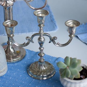 Può includere: Candelabri d'argento con più bracci, appoggiati su una tovaglia blu. I candelabri presentano dettagli ornati. È visibile anche un piccolo vaso bianco con una pianta succulenta.