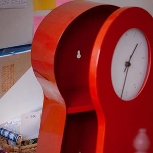 May include: A glossy red wall-mounted shelf unit with an integrated circular clock face on the right side. The unit features a keyhole-shaped silhouette with two open storage compartments and two visible keyhole mounting slots on the back panel.