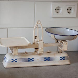 Könnte beinhalten: Eine Küchenwaage im Vintage-Stil mit einem cremefarbenen Metallsockel, der mit blauen horizontalen Streifen und quadratischen Akzenten verziert ist. Sie besitzt eine flache quadratische Platte links und eine abnehmbare, flache, weiße ovale Schale.