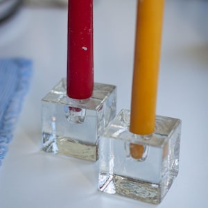 Può includere: Due portacandele a cubo di vetro con candele. Una candela è rossa, l'altra è gialla. I portacandele sono trasparenti e poggiano su una superficie bianca. Le candele sono alte e cilindriche.