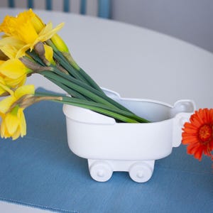 Op de afbeelding: Een witte keramische vaas in de vorm van een vintage kinderwagen met vier kleine wieltjes. De vaas is gevuld met een boeket felgele narcissen en een enkele oranje gerbera, waarbij de groene stelen zichtbaar zijn in de witte houder.