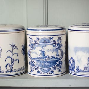 Op de afbeelding: Drie witte keramische potten met blauwe decoratieve patronen. De middelste pot toont een windmolenscène in een sierlijke lijst. De andere twee potten hebben bloemen- en landschapspatronen. De potten zijn cilindrisch met bijpassende deksels.