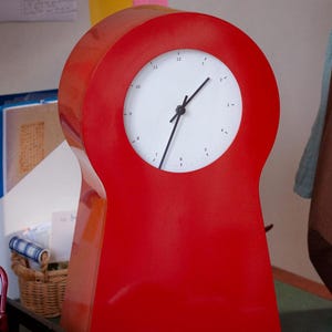 May include: A vibrant red desk clock with a minimalist design. The clock features a white face with black hands and a distinctive, rounded shape. It stands on a flat base. The clock is approximately 25 cm tall.