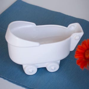 Op de afbeelding: Een witte keramische houder in de vorm van een kinderwagen met een gladde, glanzende afwerking. Het object heeft een afgeronde kap, een diepe bak en vier kleine, gevormde wielen aan de basis, ontworpen als decoratief voorwerp.
