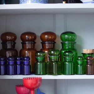 Könnte beinhalten: Ein Regal mit einer Sammlung von Glasflaschen in verschiedenen Farben und Formen. Die Flaschen sind in Reihen angeordnet und hauptsächlich braun, grün und blau. Einige der Flaschen haben Korkstopfen.