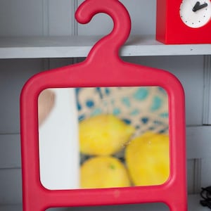 Op de afbeelding: Een felrode spiegel in de vorm van een kleerhanger, met een rechthoekig spiegeloppervlak. De reflectie toont vage gele citroenen. Een rode klok in de vorm van een huis is zichtbaar op de achtergrond.