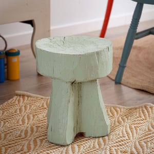May include: A small, round, light mint green wooden stool with a thick, flat top and a sturdy, carved base. The wood surface shows visible grain, natural cracks, and a weathered, matte paint finish, highlighting its solid, hand-hewn construction.