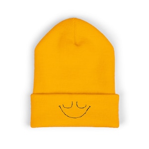 May include: Mustard yellow beanie hat with a black embroidered smiley face on the folded brim. The hat is made of a soft knit material, a casual accessory.