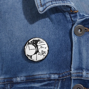 May include: A black and white round button pin featuring a cartoon character with a clock and the letters "RO". The pin is attached to a blue denim jacket.